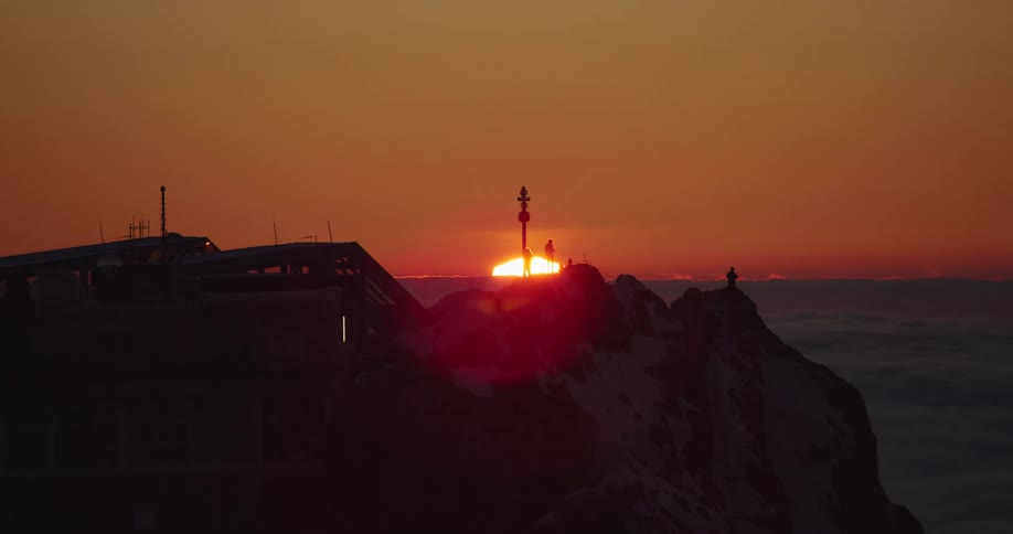 People stand on a cliff overlooking a sea of clouds as the sun begins to peak over the horizon