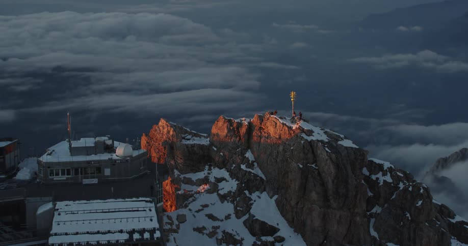 Aerial view of a cableway station atop of a snow covered mountain range over the clouds at sunset