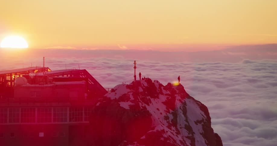 People standing atop a snow covered mountain peak near a cable car station over the clouds at sunset