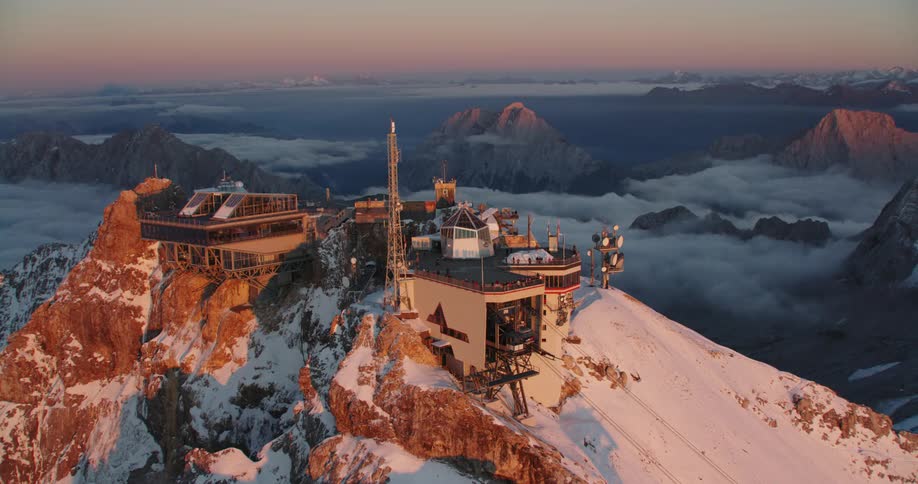 Aerial view of a cableway station atop of a snow covered mountain range over the clouds at sunset