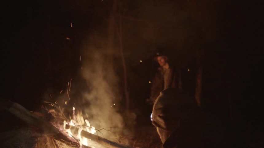 Two men near a roaring campfire and one is holding a cell phone taking a picture