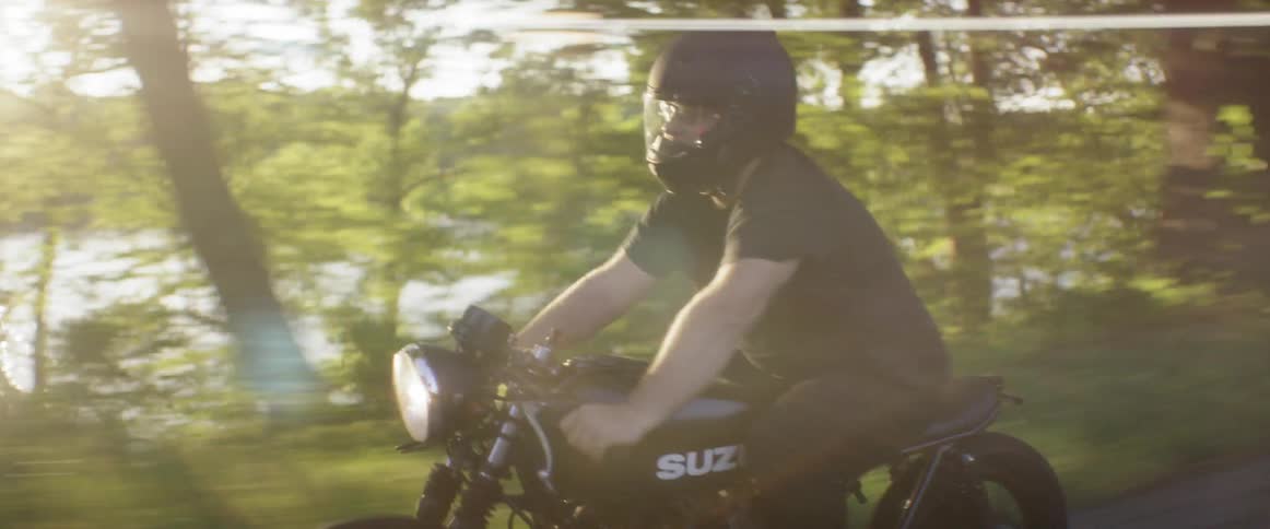 A man on a motorcycle speeds along a road in a forest near a lake