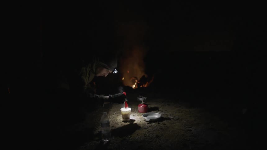 A man wearing a headlamp squatting down in a campsite stirring a cup of food