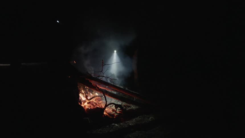 Two men wearing headlamps stoke a campfire and tend to pots