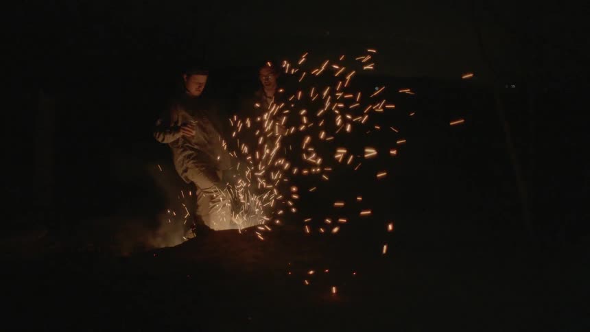 Two people are drinking by a campfire one man begins kicking the flames