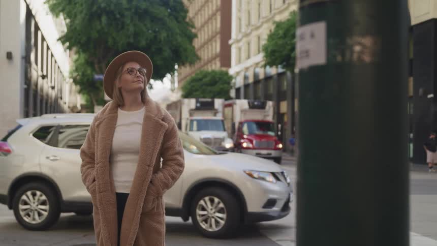 Blonde woman with a brown top coat and a brown hat stands and looks up at tall buildings in the street