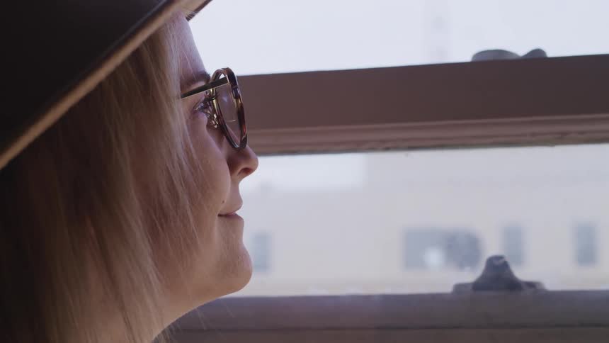 Close up of a womans face wearing glasses and a hat looking out of a window