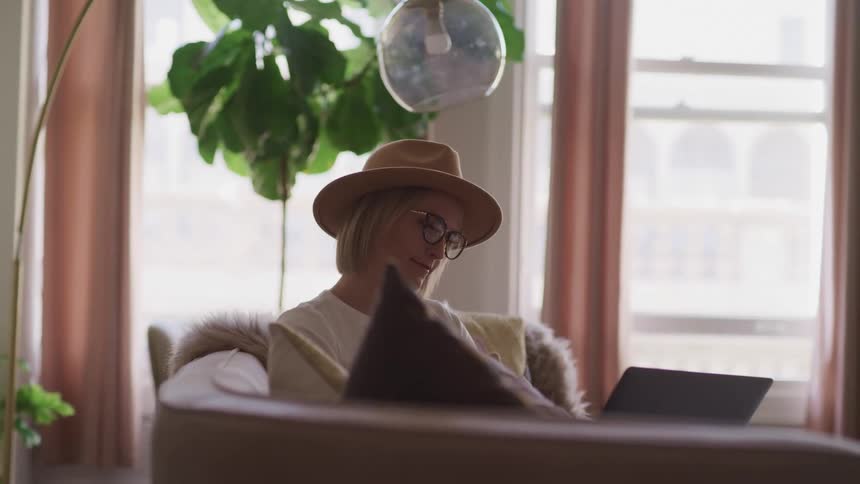 Blonde woman with a brown hat glasses and a white shirt sits on a couch and uses a laptop in the living room