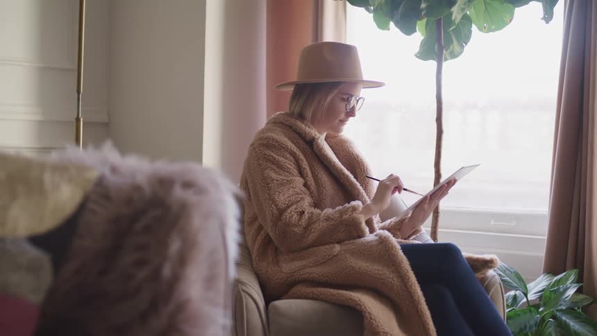 Woman wearing a fur coat and a hat draws on paper with a small brush while sitting on the living room