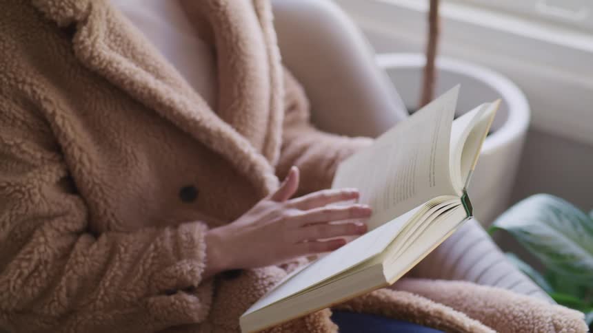 Woman with a brown overcoat sits in an armchair and turns over the page of a book