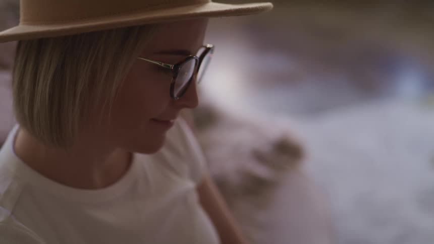 Blonde woman with a brown hat a white t shirt and glasses looks down