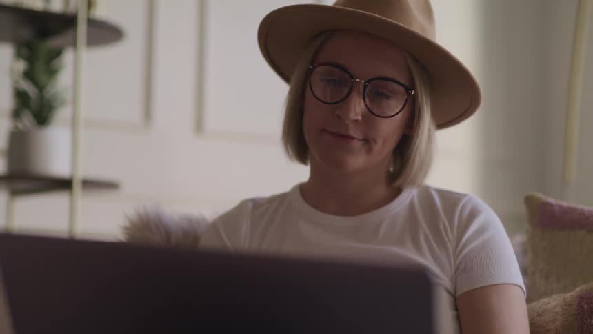 Blonde woman with glasses a brown hat sits on a couch and uses a laptop in the living room
