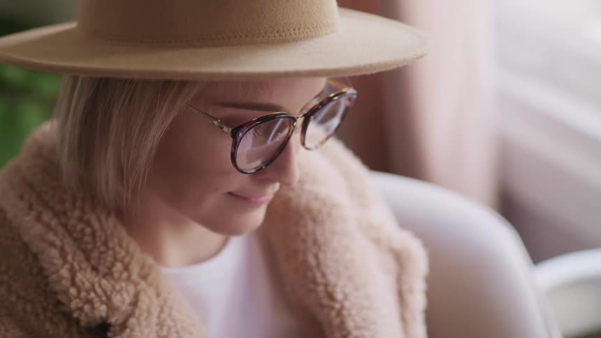 Woman with a brown hat glasses and an overcoat sits in an armchair and looks down