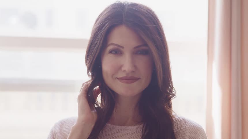 Woman with wavy hair and makeup faces the camera with eye contact in a room