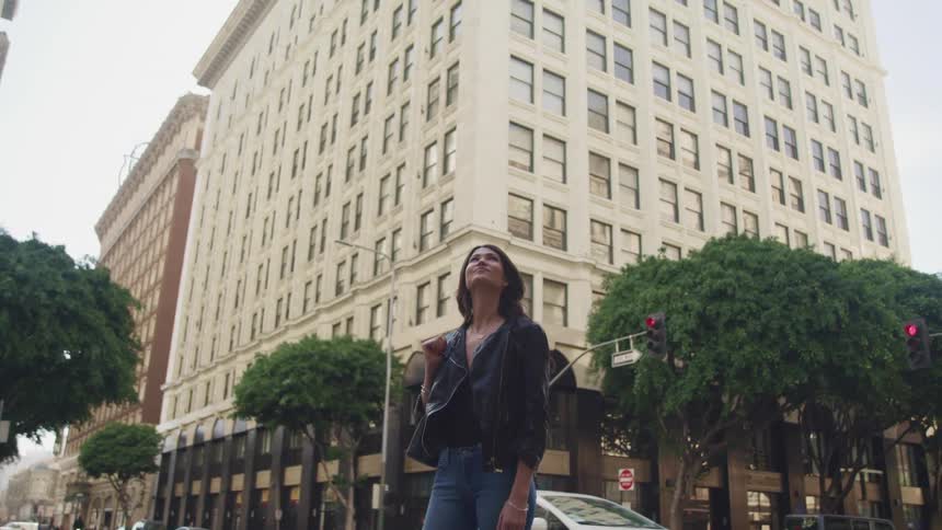 A young woman stands near a towering building while looking upwards