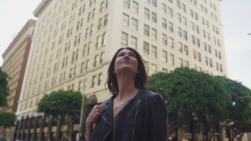 A woman gazes up at building as she stands on the sidewalk of a metropolis during the day