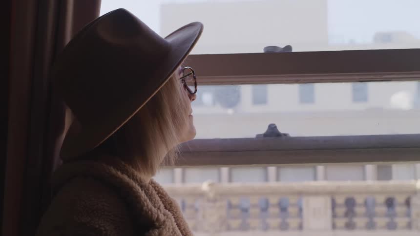 Side view of a blonde woman with a hat and glasses looking out the apartment window