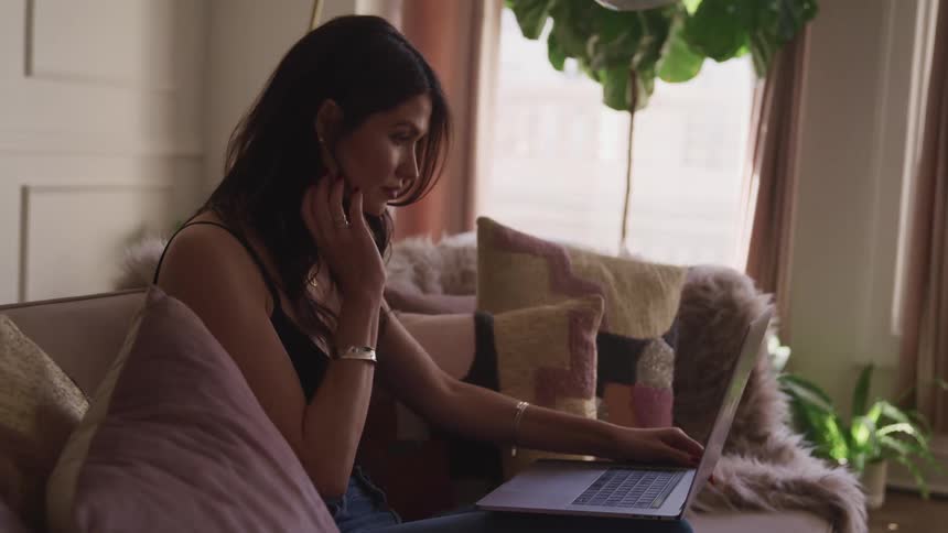 A woman touches her hair as she sits on her couch and uses a laptop
