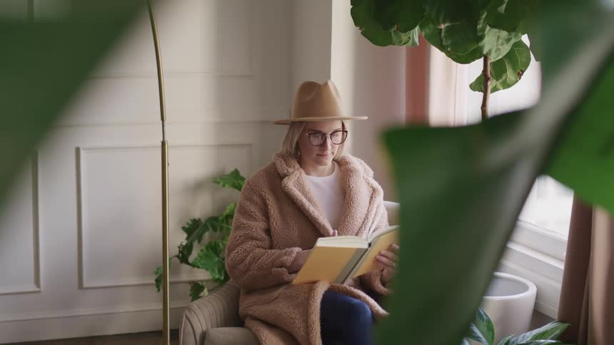Blonde woman with glasses and a brown hat and overcoat sits in an armchair and leafs through a book in a room