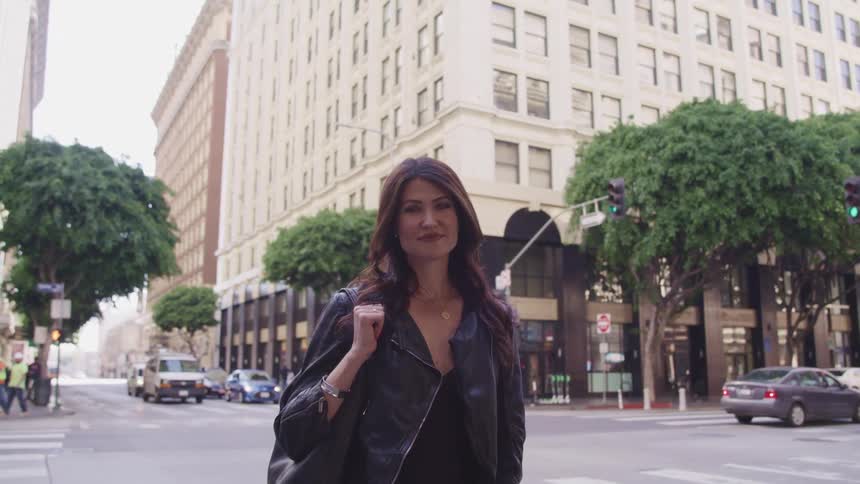 A woman stands on a sidewalk outside a large city building and intersection