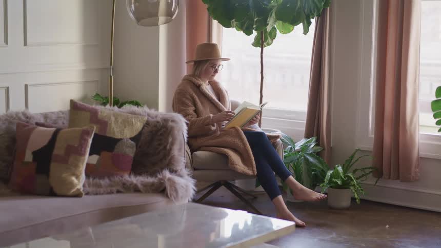 Blonde woman with a brown hat and over coat sits in an armchair and reads a book in the living room