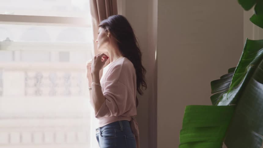 Woman with wavy hair pink sweater and jeans looks out the apartment windows