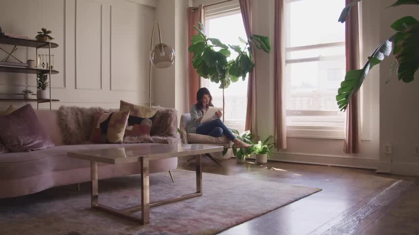 Woman sits in chair near windows in living room reading