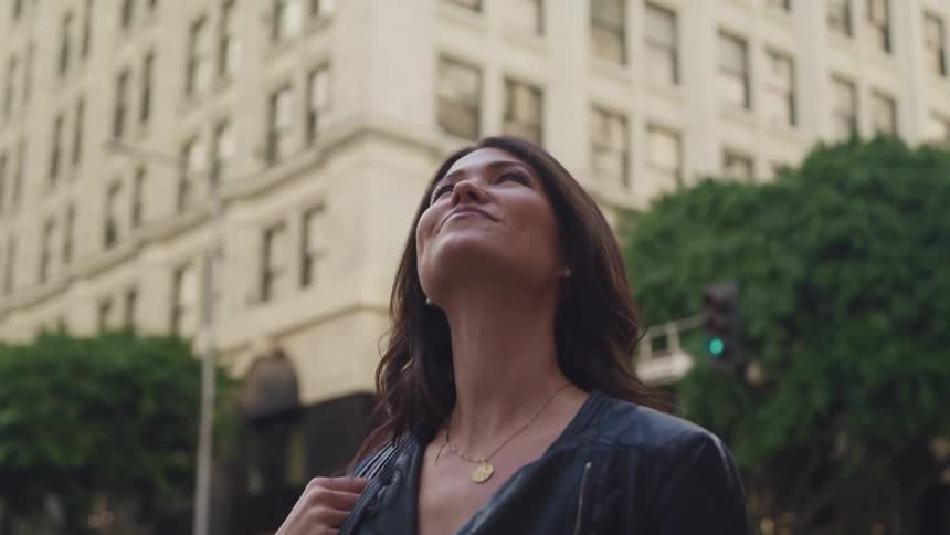 Woman with wavy hair and a black leather jacket looks up at tall buildings in city street