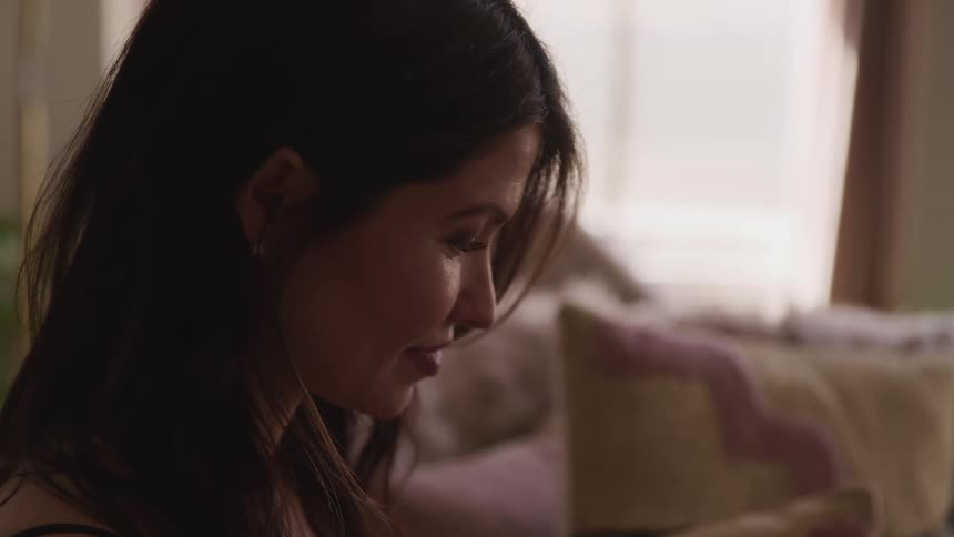 Side view of a woman with black wavy hair sitting on a sofa and looking down in a room