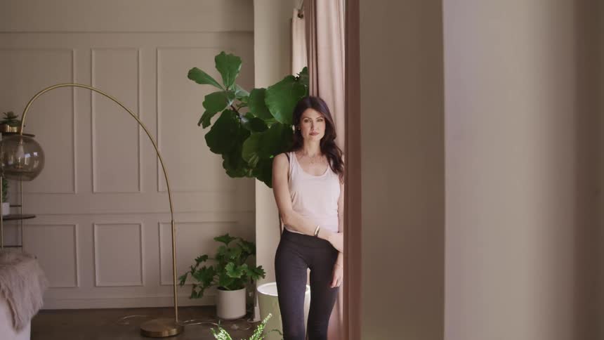 Woman with black wavy hair standing by windows smiles and looks at the camera