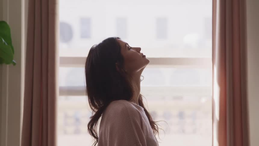 Side view of a woman with dark wavy hair looking up at ceiling and closing eyes next to an apartment window