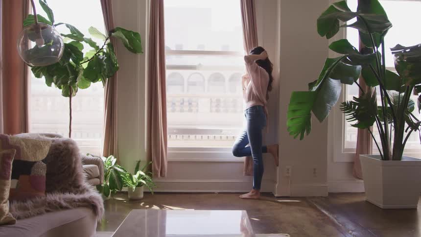 Woman stands at a window and looks outside while she runs her fingers through her long hair