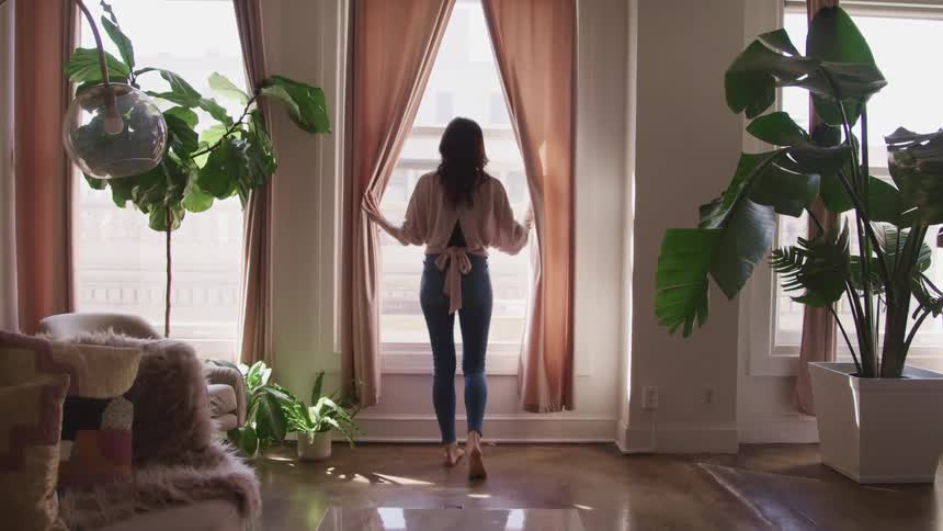 A woman at home parts the curtains and looks outside