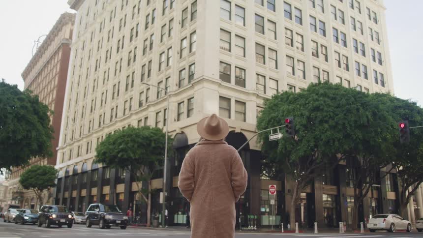 Woman with a brown hat and overcoat stands and looks at a white big building in the street