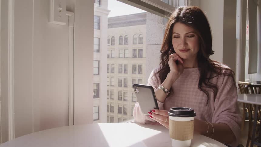 Woman with black wavy hair and a pink sweater sits at a table and uses a smart phone in a cafe