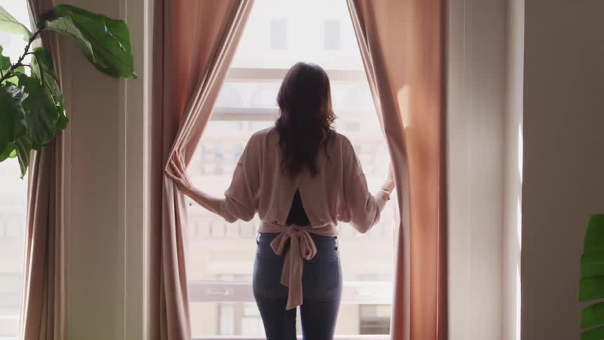 A woman approaches a tall window and separates the curtains with her hands before gazing outside
