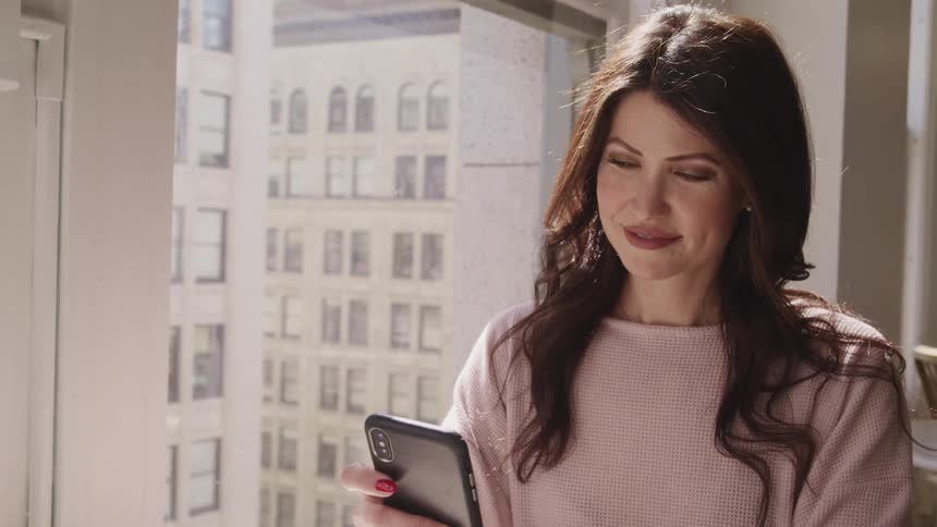 Middle aged woman with wavy black hair and a pink sweater stands next a window and uses a smartphone in an apartment