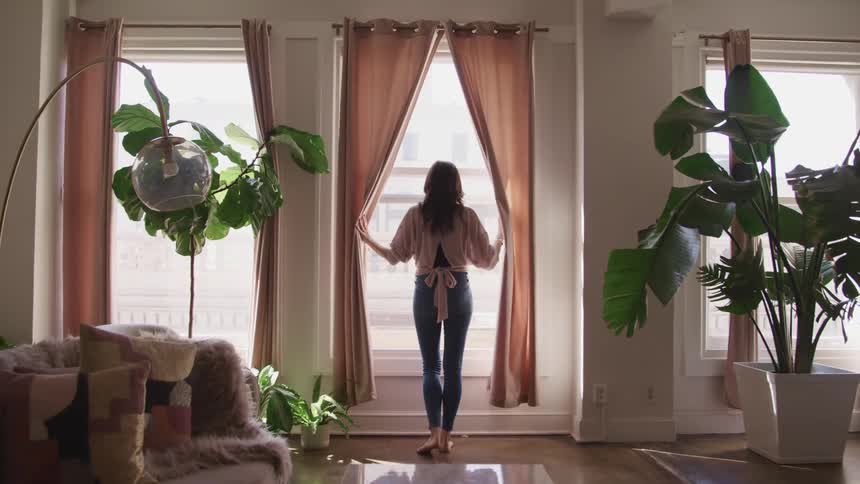 A woman walks over to a window in her apartment and pulls the pink curtains apart to look outside