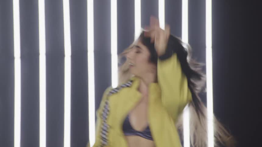 A woman dances in front of a lighted wall in a yellow track jacket and sports bra