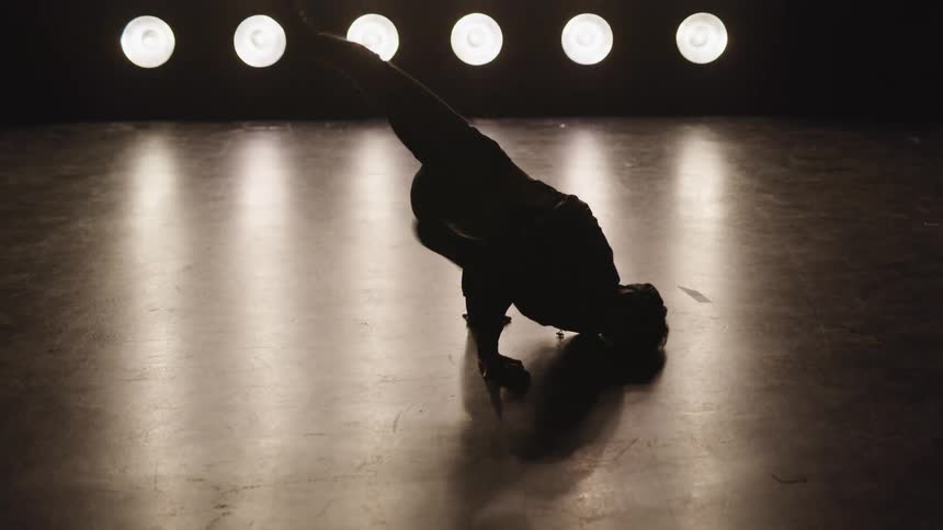 A man walks on his hands and performs break dancing poses in front of large light fixture