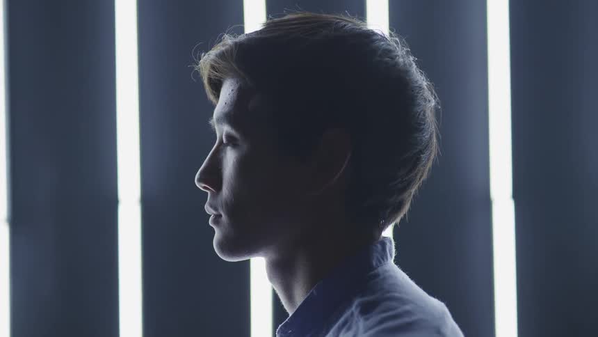 A young asian man standing in a front of a wall with fluorescent lights mounted vertically on it slowly raises his head
