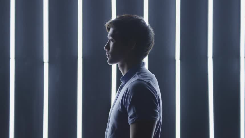 A young looks up as he stands in profile against a wall with vertical lines of light