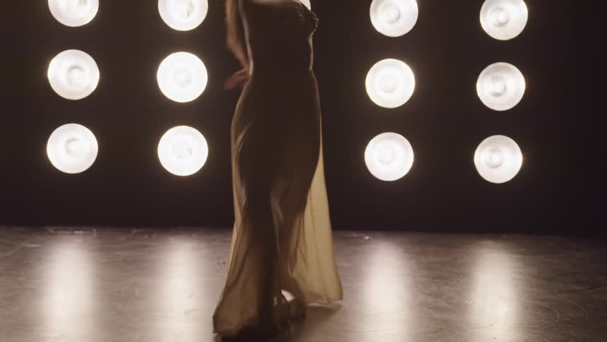 A woman in a long sheer dress dances in a studio with dozens of circular lights on a wall