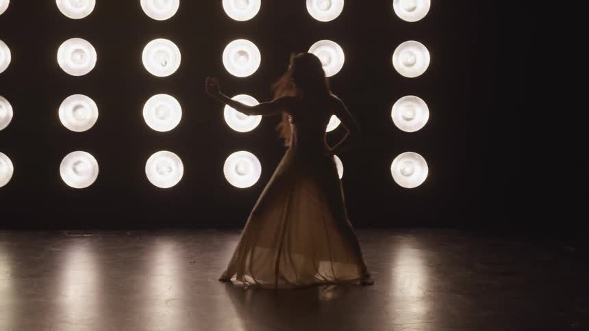 Woman in a long transparent dress dances in front of a wall of floodlights on stage