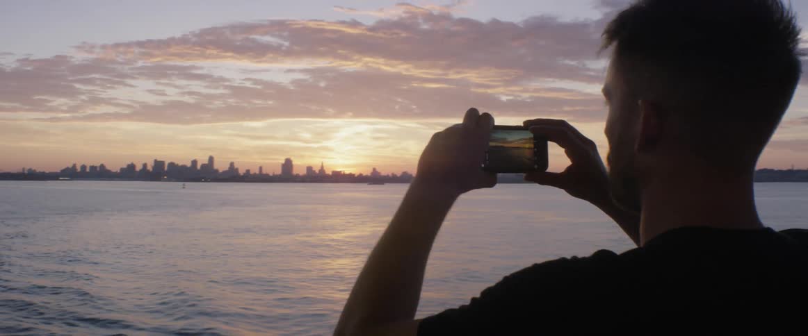 A man takes a picture of a cityscape from a boat at sunset