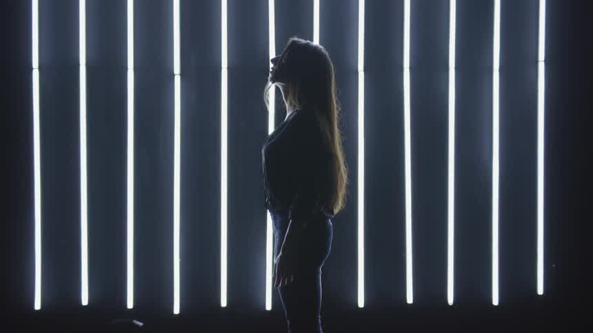 Woman stands in darkroom backlit by parallel row of vertical fluorescent lights