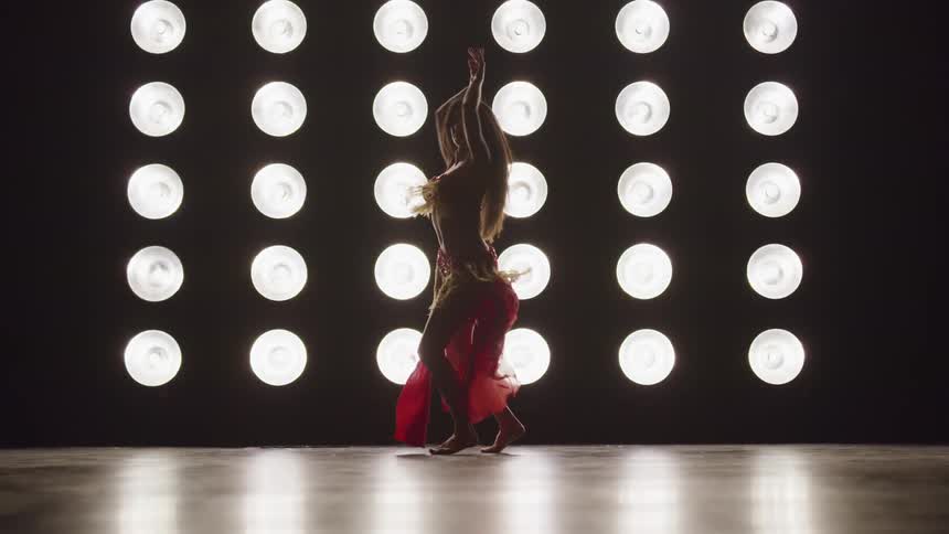 A woman does a belly dance on a stage in front of set of bright stage lights