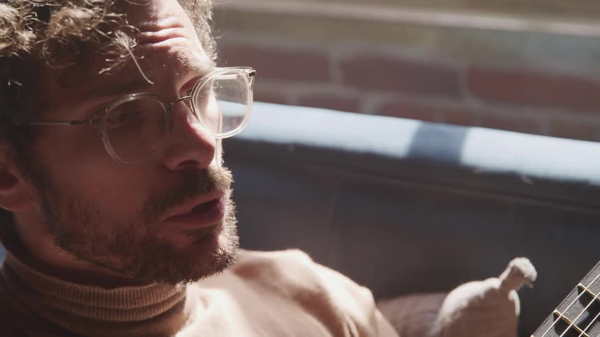 Man with a beard glasses and a brown turtleneck sweater plays a guitar while singing a song