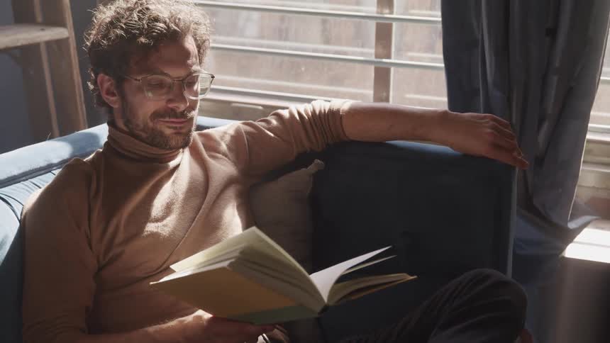 Man with brown turtleneck sweater a beard and glasses sits on a sofa reads a book in the living room