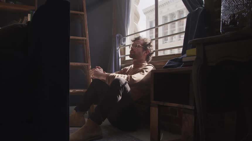 Man with a brown turtleneck sweater sits and leans against the wall under a window in a room